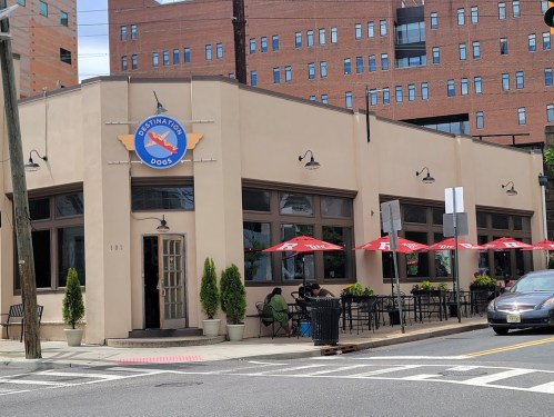 Exterior of a restaurant called "Destination Dogs," featuring outdoor seating and a modern urban backdrop.