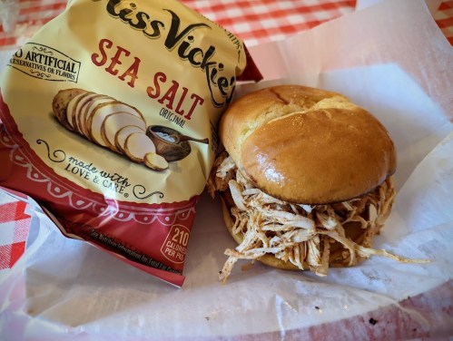 A pulled chicken sandwich on a bun next to a bag of Miss Vickie's sea salt chips on a checkered tablecloth.