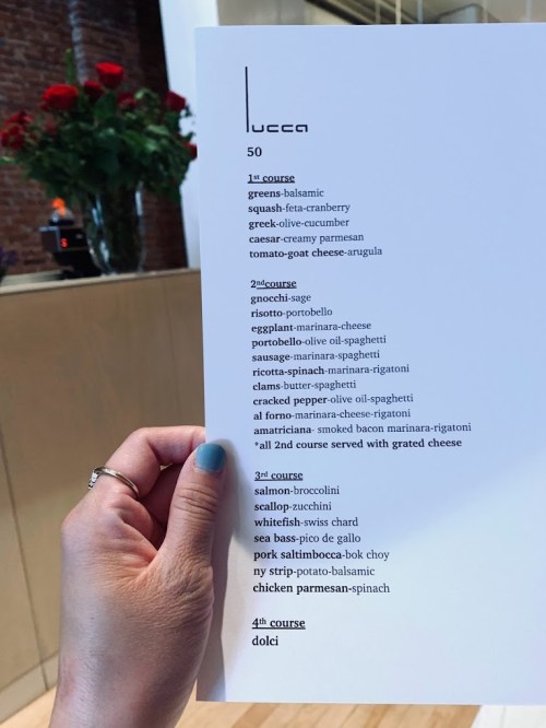 A hand holding a menu from Lucca featuring four courses with various Italian dishes and ingredients listed.