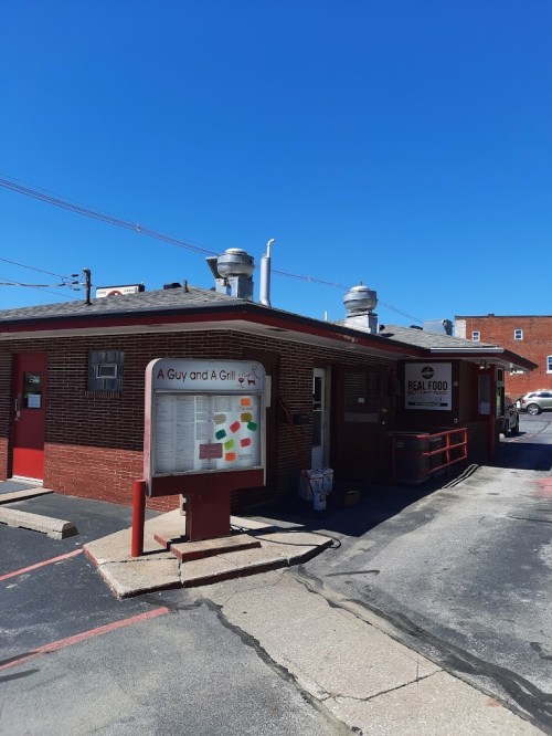 A red brick building with a sign reading "A Guy and A Grill" and a menu board outside, under a clear blue sky.