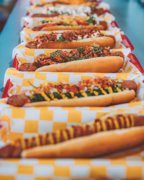 A row of colorful hot dogs topped with various ingredients, served in red and white checkered baskets.