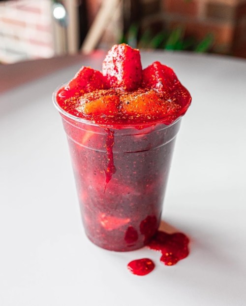 A clear cup filled with vibrant red fruit, topped with diced strawberries and a sprinkle of seasoning, with some juice spilling out.
