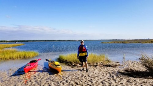 A person stands on a sandy shore beside two kayaks, with a calm waterway and grassy areas in the background.