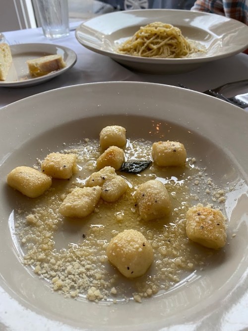 A plate of gnocchi in a light sauce with grated cheese, alongside a bowl of spaghetti in the background.