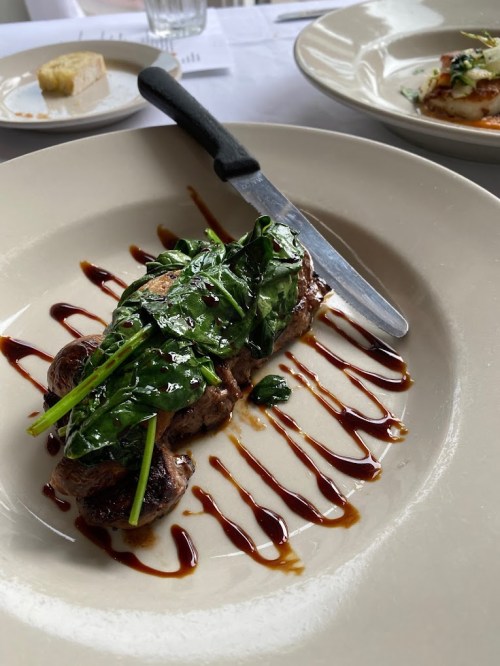A plated dish featuring a piece of meat topped with sautéed spinach and drizzled with sauce, accompanied by a knife.