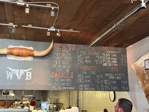 Menu board displaying meats, sauces, platters, and sandwiches with prices at a barbecue restaurant.