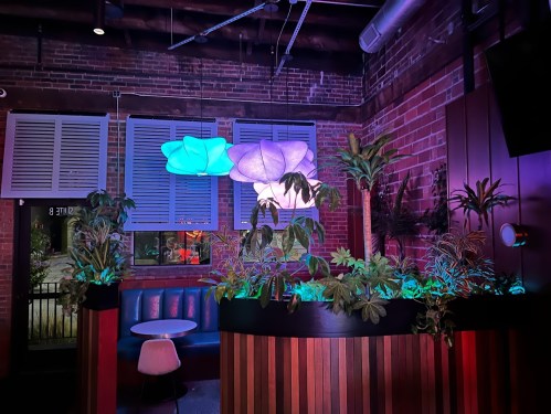 A stylish bar interior with colorful lighting, plants, and unique pendant lamps against a brick wall.