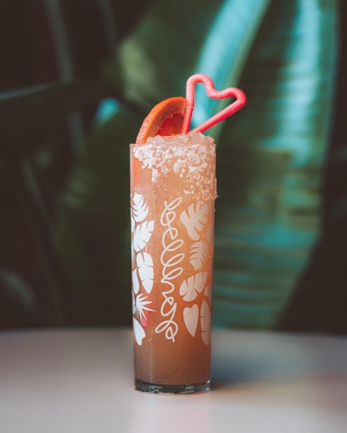 A tall cocktail glass with a decorative design, garnished with a slice of orange and two pink straws, set against green foliage.