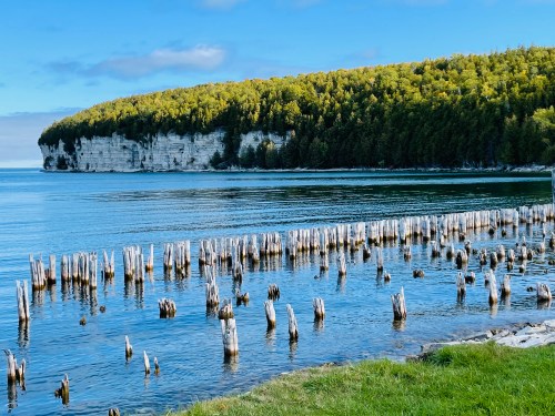 A serene lake scene with wooden posts emerging from the water, surrounded by lush green hills and a clear blue sky.
