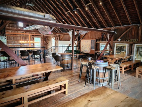 A rustic barn interior with wooden tables, bar stools, and a cozy atmosphere, featuring exposed beams and warm lighting.