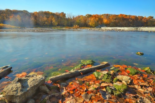 A serene river scene with colorful autumn leaves, rocks, and trees reflecting in the calm water under a clear blue sky.