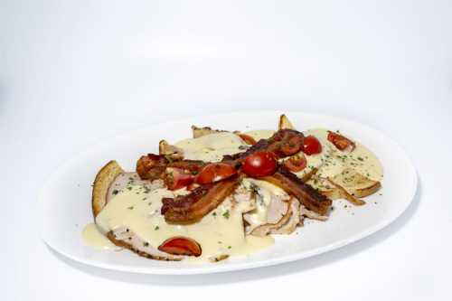 A plate of sliced turkey topped with bacon, cherry tomatoes, and a creamy sauce, served with crispy potato wedges.