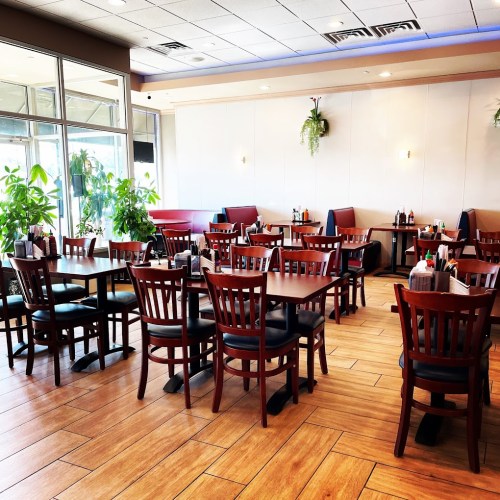 A bright, empty restaurant with wooden tables and chairs, plants, and booths along the walls.