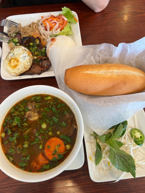 A bowl of soup with vegetables and herbs, a plate of rice, grilled meat, an egg, and a fresh baguette on a table.