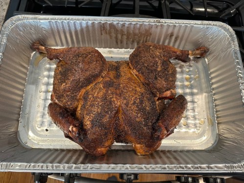 A seasoned, roasted chicken in a foil pan, resting on a stovetop.