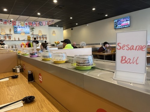 A restaurant interior with a display of colorful desserts, including a sign for "Sesame Ball," and diners in the background.