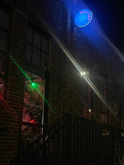A brick building at night with a glowing blue sign and colorful lights visible through the windows.