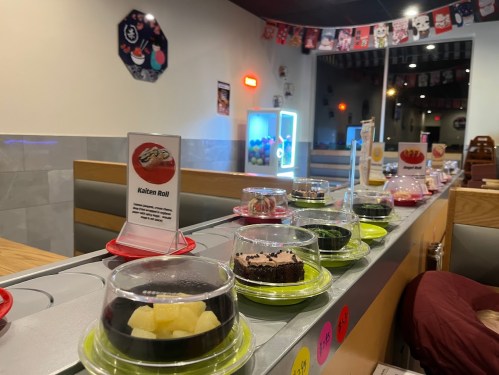 A conveyor belt displays various colorful desserts in small containers at a restaurant, with a sign for "Kaiten Roll."