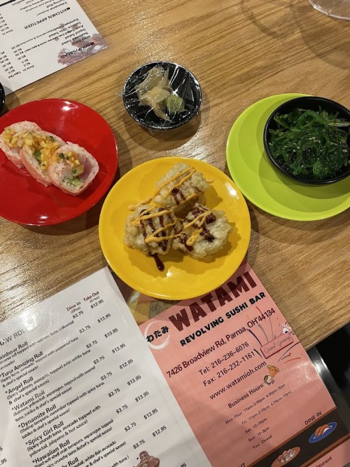 A table with sushi rolls, a yellow plate of tempura, seaweed salad, and a menu from Watami Sushi Bar.