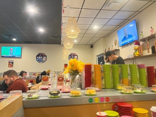 A vibrant restaurant interior with colorful plates stacked, a TV showing news, and customers enjoying their meals.