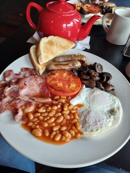 A hearty breakfast plate with bacon, sausages, eggs, baked beans, grilled tomato, mushrooms, and toast, alongside a red teapot.