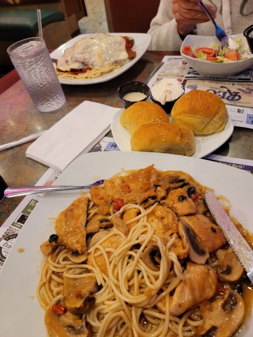 A table setting with a plate of chicken pasta, rolls, a salad, and a dish of baked chicken with cheese.