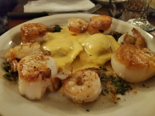 A plate of seafood featuring shrimp, scallops, and ravioli in a creamy sauce, garnished with herbs.