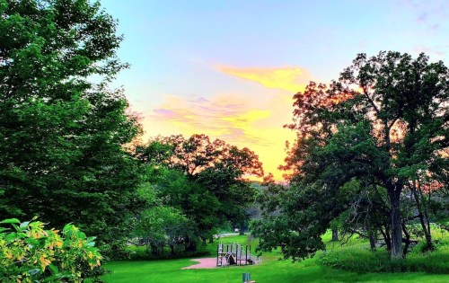 A vibrant sunset over a green park, with trees framing a playground in the foreground.