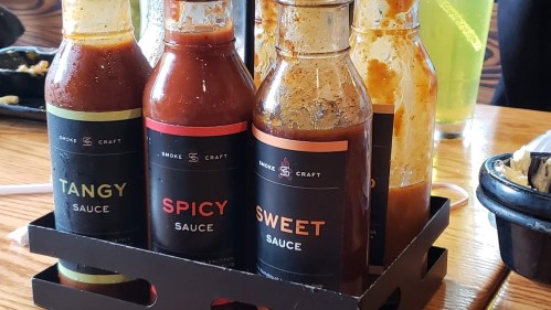 A variety of sauce bottles labeled Tangy, Spicy, and Sweet, arranged in a holder on a wooden table.
