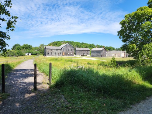 A scenic view of old wooden buildings surrounded by green fields and trees under a blue sky with scattered clouds.