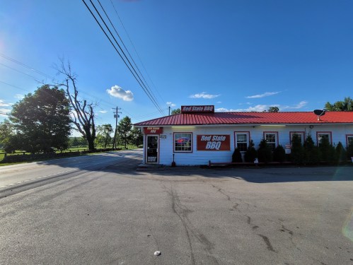 A BBQ restaurant with a red roof, surrounded by trees and a clear blue sky, located at a road intersection.