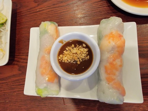 Two fresh spring rolls with shrimp on a plate, accompanied by a small bowl of peanut sauce topped with crushed peanuts.