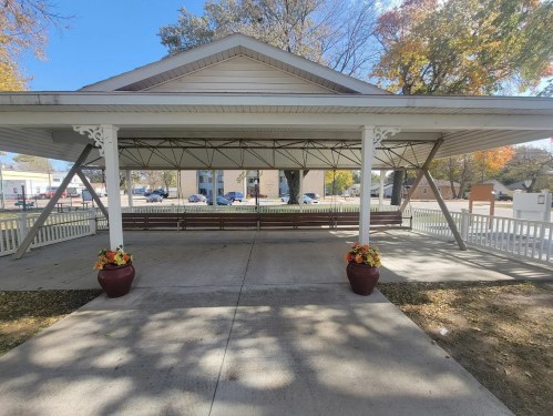 A covered pavilion with a wooden bench, surrounded by flower pots, set in a park with trees and a clear blue sky.