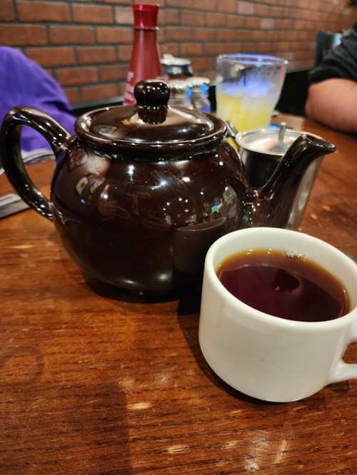 A black teapot sits on a wooden table next to a white cup filled with tea.