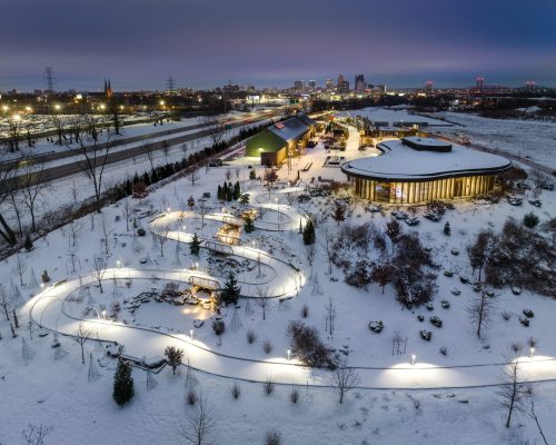 Aerial view of a snowy landscape with winding paths, illuminated gardens, and a modern building against a city skyline.