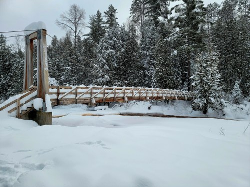 A snow-covered wooden suspension bridge spans a frozen river, surrounded by tall evergreen trees in a winter landscape.