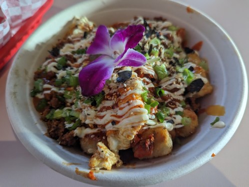 A bowl of loaded nachos topped with green onions, sesame seeds, drizzled sauce, and a purple flower garnish.