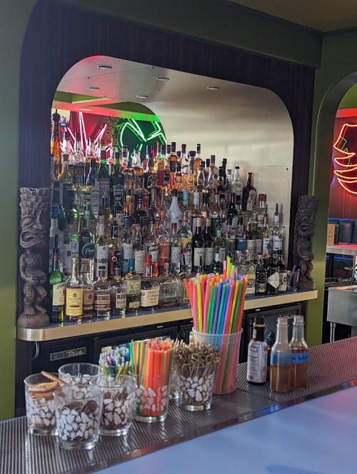 A vibrant bar with a variety of liquor bottles, colorful straws, and decorative tiki elements. Neon lights in the background.
