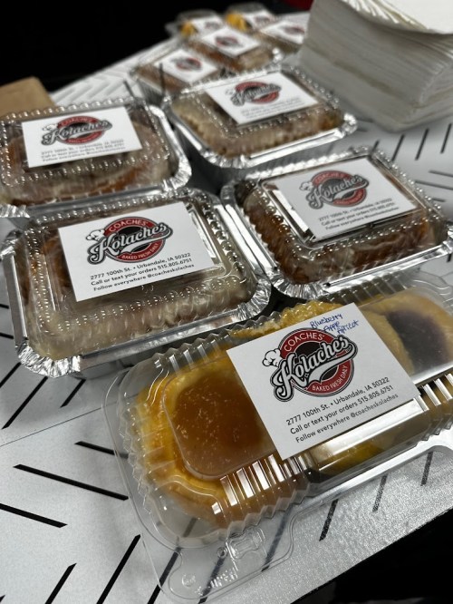 A variety of kolaches in clear containers, labeled with a logo and address, arranged on a table.