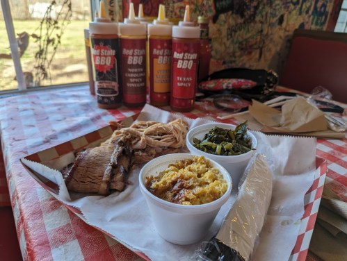 A plate of BBQ with pulled pork, collard greens, and cornbread, alongside bottles of BBQ sauce on a checkered tablecloth.