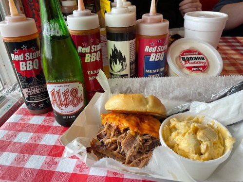 A plate with pulled pork sandwich, mac and cheese, a bottle of soda, and various BBQ sauces in the background.