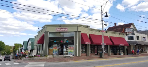A storefront with red awnings and large windows, surrounded by trees and a blue sky with clouds.