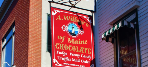 Sign for A. Wilbur's of Maine Chocolate, featuring fudge, penny candy, and truffles, with a red background and lighthouse logo.