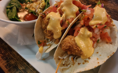Two tacos filled with fried chicken, topped with cheese sauce, tomatoes, and onions, served with a side salad.