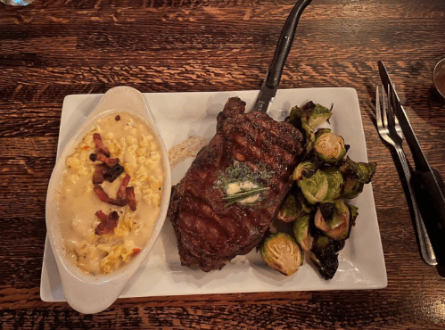 A plate featuring a grilled steak with butter, creamy corn casserole, and roasted Brussels sprouts on a wooden table.