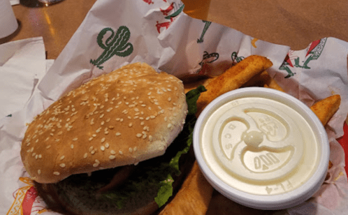 A sesame seed bun burger with lettuce, served with fries and a cup of dipping sauce on a colorful paper liner.