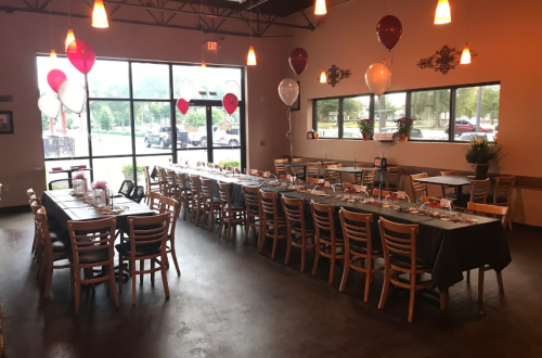 A decorated restaurant interior with a long table set for a gathering, featuring balloons and a view of the outside.