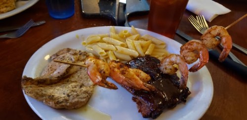 A plate with grilled shrimp skewers, pasta, and two types of meat: chicken and steak.