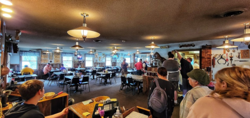 A busy restaurant interior with patrons waiting in line and tables set for dining. Warm lighting and a cozy atmosphere.