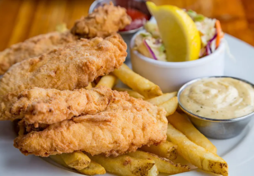 A plate of crispy fried fish fillets served with French fries, coleslaw, lemon, and dipping sauces.